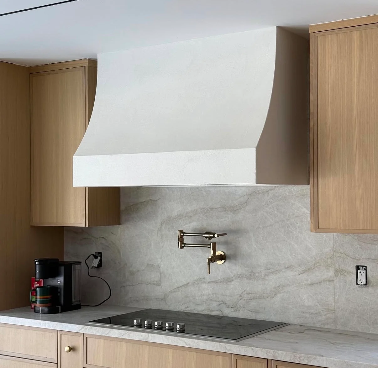 Pale microcement finish on a sculpted kitchen range hood between natural wood cabinets above a stone backsplash
